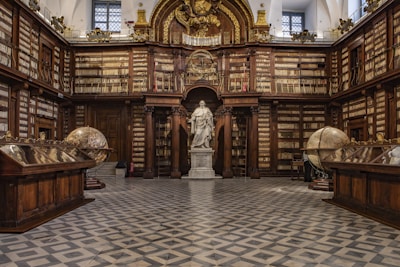 A grand, classical library interior featuring ornate wooden bookshelves filled with books that line the walls. At the center stands a prominent statue of a historical figure surrounded by architectural details like columns and arches. Two large, antique globes are positioned on either side of the room, and a checkered floor adds to the elegant ambiance.