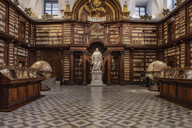 A grand, classical library interior featuring ornate wooden bookshelves filled with books that line the walls. At the center stands a prominent statue of a historical figure surrounded by architectural details like columns and arches. Two large, antique globes are positioned on either side of the room, and a checkered floor adds to the elegant ambiance.