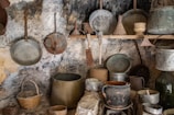 A rustic kitchen scene featuring various old, weathered cooking utensils and containers. Metal pans, pots, and sieves with a vintage appearance hang from hooks on a rough, textured stone wall. A woven basket sits on a lower shelf, and larger pots are arranged on rocks and surfaces, adding to the historical ambiance.