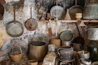 A rustic kitchen scene featuring various old, weathered cooking utensils and containers. Metal pans, pots, and sieves with a vintage appearance hang from hooks on a rough, textured stone wall. A woven basket sits on a lower shelf, and larger pots are arranged on rocks and surfaces, adding to the historical ambiance.