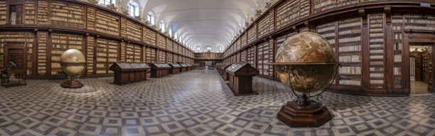 A grand, historical library with tall wooden bookshelves that extend to the ceiling and line both sides of a long aisle. The floor is covered with a geometric patterned tile, and the ceiling is arched. Large antique globes are placed on stands within the center of the room, which add to the scholarly and aged atmosphere.