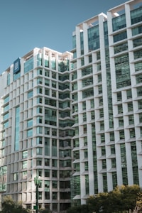 The image features a modern high-rise building with a geometric facade design, characterized by white structures and glass windows. The building reflects a clean, contemporary architectural style with multiple stories and a distinct vertical pattern. A clear sky serves as the background, enhancing the building's sleek appearance.