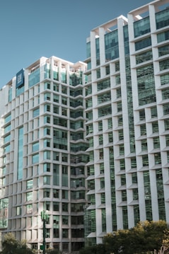 The image features a modern high-rise building with a geometric facade design, characterized by white structures and glass windows. The building reflects a clean, contemporary architectural style with multiple stories and a distinct vertical pattern. A clear sky serves as the background, enhancing the building's sleek appearance.