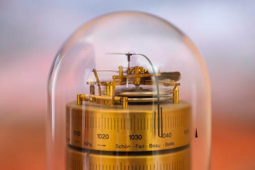 A brass barometer encased in a transparent dome is centered in the image. The barometer features intricate mechanical components and detailed measurement marks, with numbers ranging from 1000 to 1040 and words like 'Schön' and 'Beau.' The background is softly blurred with warm tones.