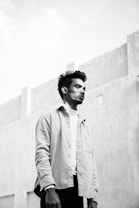 A black and white portrait of a man looking into the distance with a calm expression. The background features a textured concrete wall and a clear sky, creating a minimalist and serene setting. The man is wearing a light-colored shirt over a white turtleneck, adding a contrasting element to the monochrome palette.
