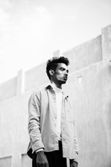 A black and white portrait of a man looking into the distance with a calm expression. The background features a textured concrete wall and a clear sky, creating a minimalist and serene setting. The man is wearing a light-colored shirt over a white turtleneck, adding a contrasting element to the monochrome palette.