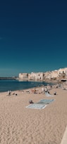 A sandy beach with several towels and beachgoers scattered across it, set against a backdrop of calm blue waters and a clear sky. In the distance, a coastal town with charming, light-colored buildings hugs the shoreline.