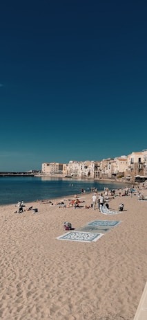 A sandy beach with several towels and beachgoers scattered across it, set against a backdrop of calm blue waters and a clear sky. In the distance, a coastal town with charming, light-colored buildings hugs the shoreline.
