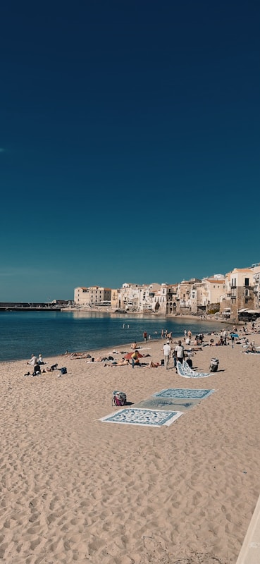 A sandy beach with several towels and beachgoers scattered across it, set against a backdrop of calm blue waters and a clear sky. In the distance, a coastal town with charming, light-colored buildings hugs the shoreline.