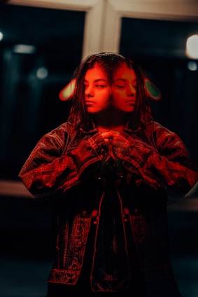 A person in a denim jacket stands with a reflective, surreal double exposure effect, creating an intriguing visual of two faces. The lighting casts a red hue over the image, enhancing the mysterious atmosphere.