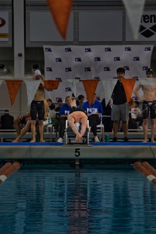 A swimmer is on a starting block poised to dive into an indoor pool with clear blue water. Surrounding the swimmer are other competitors and officials, some wearing sports attire and swim caps. Orange and white triangular flags hang above, and a banner with a logo is displayed in the background.