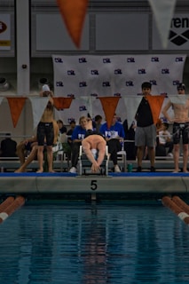A swimmer is on a starting block poised to dive into an indoor pool with clear blue water. Surrounding the swimmer are other competitors and officials, some wearing sports attire and swim caps. Orange and white triangular flags hang above, and a banner with a logo is displayed in the background.