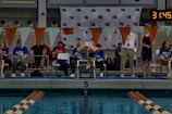 A swimmer is poised on a diving block, ready to dive into the pool, surrounded by a group of officials and spectators. The setting is an indoor swimming competition with timing equipment visible in the background. Flags are strung across the pool, and the water reflects the surrounding environment.