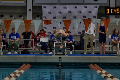 A swimmer is poised on a diving block, ready to dive into the pool, surrounded by a group of officials and spectators. The setting is an indoor swimming competition with timing equipment visible in the background. Flags are strung across the pool, and the water reflects the surrounding environment.