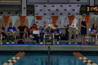 A group of CNCA swimming officials attentively observing a swim meet, clipboard in hand, by the poolside.