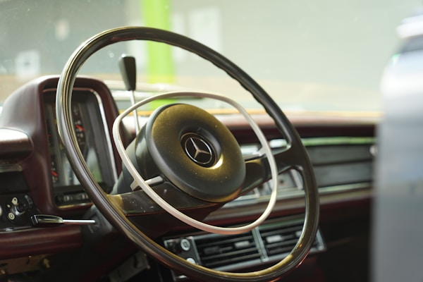 The image features the interior of a vintage car, focusing on the steering wheel with a classic Mercedes-Benz logo in the center. The dashboard shows a retro design with analog dials and wood accents. The overall atmosphere is nostalgic, emphasizing the craftsmanship and style of the era.