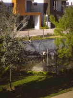 A residential area with modern architectural elements, featuring a building with a mix of orange and white tones. The foreground includes a small pond or water feature with a pipe and surrounded by a metal fence. Lush greenery and trees add to the tranquil outdoor atmosphere.