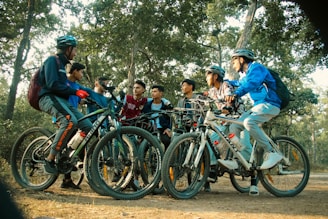 A group of young people are gathered in a wooded area, each with a bicycle. They appear to be engaged in conversation and are dressed in casual, sporty clothing including helmets. The setting is a forest with tall trees and a dirt trail, suggesting an outdoor adventure or cycling trip.