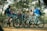 A group of young people are gathered in a wooded area, each with a bicycle. They appear to be engaged in conversation and are dressed in casual, sporty clothing including helmets. The setting is a forest with tall trees and a dirt trail, suggesting an outdoor adventure or cycling trip.