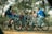 A group of young people are gathered in a wooded area, each with a bicycle. They appear to be engaged in conversation and are dressed in casual, sporty clothing including helmets. The setting is a forest with tall trees and a dirt trail, suggesting an outdoor adventure or cycling trip.