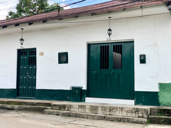 A white building features two green doors, one single and one double, with small rectangular windows near the top. Two black lanterns are fixed above each door. There is a small green box or vent to the right of the double doors. The building's lower wall section is painted green, and the roof is a muted red-brown.