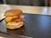 A juicy cheeseburger sits on a black tray with a toasted bun, melted cheddar cheese, a beef patty, fresh tomato slices, crisp lettuce, and creamy sauce. The background is softly blurred, creating focus on the burger.