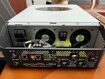 A piece of electronic equipment with multiple input and output ports is placed on a wooden surface. The device has fans for cooling, connection ports labeled with various functions, and several wires and circuit elements visible on the front panel.
