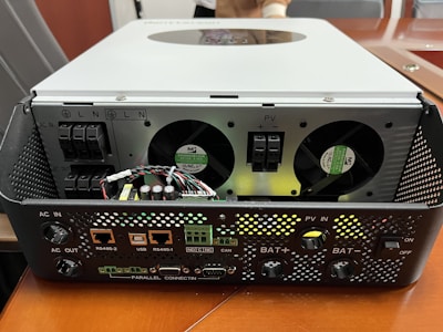 A piece of electronic equipment with multiple input and output ports is placed on a wooden surface. The device has fans for cooling, connection ports labeled with various functions, and several wires and circuit elements visible on the front panel.