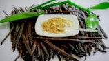 A white plate containing a pile of brown powder is placed on a surface covered in vanilla beans. A small white spoon rests on the plate. Bright green leaves add a touch of color to the composition.