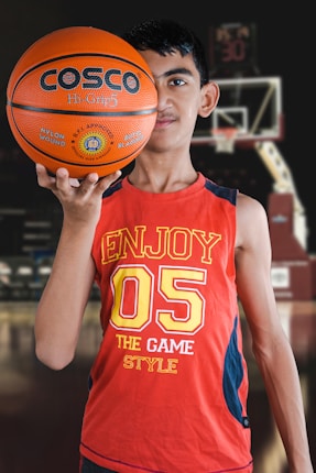 A young person holds a basketball with one hand in the foreground, wearing a red sleeveless shirt with the number 05 and the words 'Enjoy the Game Style.' The background features a basketball hoop and blurred elements that suggest an indoor basketball court.