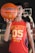 A young person holds a basketball with one hand in the foreground, wearing a red sleeveless shirt with the number 05 and the words 'Enjoy the Game Style.' The background features a basketball hoop and blurred elements that suggest an indoor basketball court.