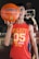 A young person holds a basketball with one hand in the foreground, wearing a red sleeveless shirt with the number 05 and the words 'Enjoy the Game Style.' The background features a basketball hoop and blurred elements that suggest an indoor basketball court.