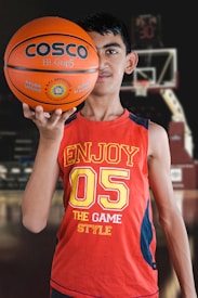 A young person holds a basketball with one hand in the foreground, wearing a red sleeveless shirt with the number 05 and the words 'Enjoy the Game Style.' The background features a basketball hoop and blurred elements that suggest an indoor basketball court.
