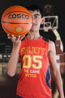 A young person holds a basketball with one hand in the foreground, wearing a red sleeveless shirt with the number 05 and the words 'Enjoy the Game Style.' The background features a basketball hoop and blurred elements that suggest an indoor basketball court.