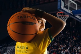 A young person is holding a basketball in front of a large crowd in a sports arena. They are wearing a yellow jersey with green trim, which has text on it, and are smiling confidently.