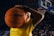 A young person is holding a basketball in front of a large crowd in a sports arena. They are wearing a yellow jersey with green trim, which has text on it, and are smiling confidently.
