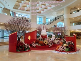 A festive indoor display features large white bunny sculptures surrounded by floral arrangements in shades of red, pink, and orange. The setting appears to be inside a spacious mall or commercial area with escalators and modern architecture. Decorative elements include red panels and cherry blossom branches, contributing to a celebratory and seasonal atmosphere.