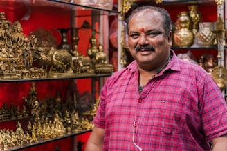 A person wearing a red plaid shirt stands next to a display of intricately designed golden brass sculptures depicting various figures and elephants. The background is a vibrant red, enhancing the richness of the displayed items.