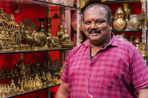 A person wearing a red plaid shirt stands next to a display of intricately designed golden brass sculptures depicting various figures and elephants. The background is a vibrant red, enhancing the richness of the displayed items.