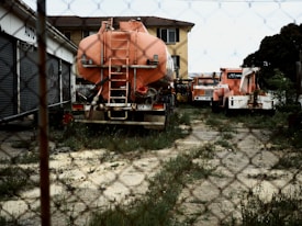 The image features a fenced-off yard with several orange heavy-duty trucks and tankers parked on an overgrown plot. On the left, there is a building with closed metal shutters and a sign partially visible that reads 'AUTO-'. In the background, there is a multi-story yellow brick building.