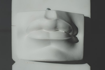A close-up view of a sculpted piece depicting the nose and lips portion of a human face, made from a smooth, light-colored material. The lighting casts soft shadows that enhance the contours and details of the sculpture.