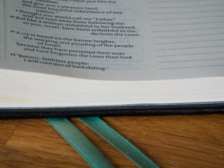 A close-up view of an open book showing a page with text printed in a column format. The page includes verse numbers on the left and a section of text highlighted. Two green ribbons are used as bookmarks, resting on a wooden surface.