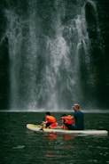 A group of people wearing bright orange life jackets paddle together on a yellow and white inflatable raft. In the background, a majestic waterfall cascades down a rugged, dark stone cliff, creating a misty spray at its base.