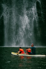 A group of people wearing bright orange life jackets paddle together on a yellow and white inflatable raft. In the background, a majestic waterfall cascades down a rugged, dark stone cliff, creating a misty spray at its base.