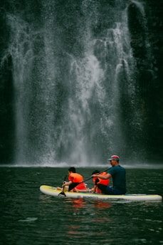 A group of people wearing bright orange life jackets paddle together on a yellow and white inflatable raft. In the background, a majestic waterfall cascades down a rugged, dark stone cliff, creating a misty spray at its base.