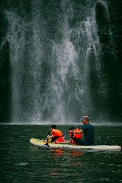 A group of people wearing bright orange life jackets paddle together on a yellow and white inflatable raft. In the background, a majestic waterfall cascades down a rugged, dark stone cliff, creating a misty spray at its base.