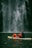 A group of people wearing bright orange life jackets paddle together on a yellow and white inflatable raft. In the background, a majestic waterfall cascades down a rugged, dark stone cliff, creating a misty spray at its base.