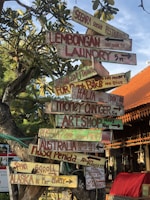 A creatively crafted signpost with multiple colorful wooden signs pointing in different directions. The signs display various whimsical and humorous messages, such as directions to distant places like Alaska and Russia, and local spots like Lembongan Laundry and a Seaside Restaurant. The background includes lush green foliage and an orange-tiled roof of a nearby building under a clear blue sky.