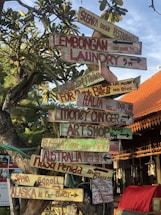 A creatively crafted signpost with multiple colorful wooden signs pointing in different directions. The signs display various whimsical and humorous messages, such as directions to distant places like Alaska and Russia, and local spots like Lembongan Laundry and a Seaside Restaurant. The background includes lush green foliage and an orange-tiled roof of a nearby building under a clear blue sky.