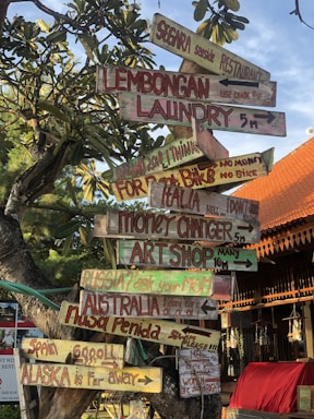 A creatively crafted signpost with multiple colorful wooden signs pointing in different directions. The signs display various whimsical and humorous messages, such as directions to distant places like Alaska and Russia, and local spots like Lembongan Laundry and a Seaside Restaurant. The background includes lush green foliage and an orange-tiled roof of a nearby building under a clear blue sky.